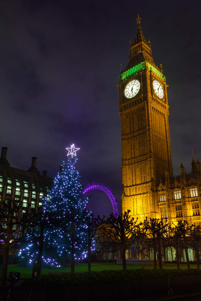 Big Ben tower and clock lit up at night, with Christmas tree in the foreground and the London Eye in the background
