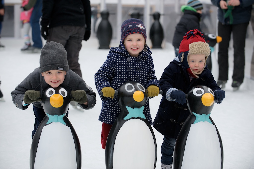 Three children skating on an ice rink pushing plastic penguins